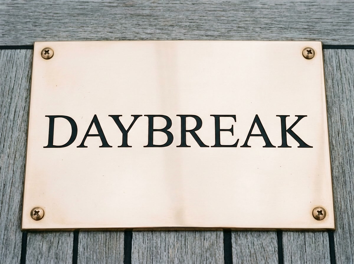 Polished bronze yacht transom plate engraved DAYBREAK, photographed straight on against weathered teak deck planking
