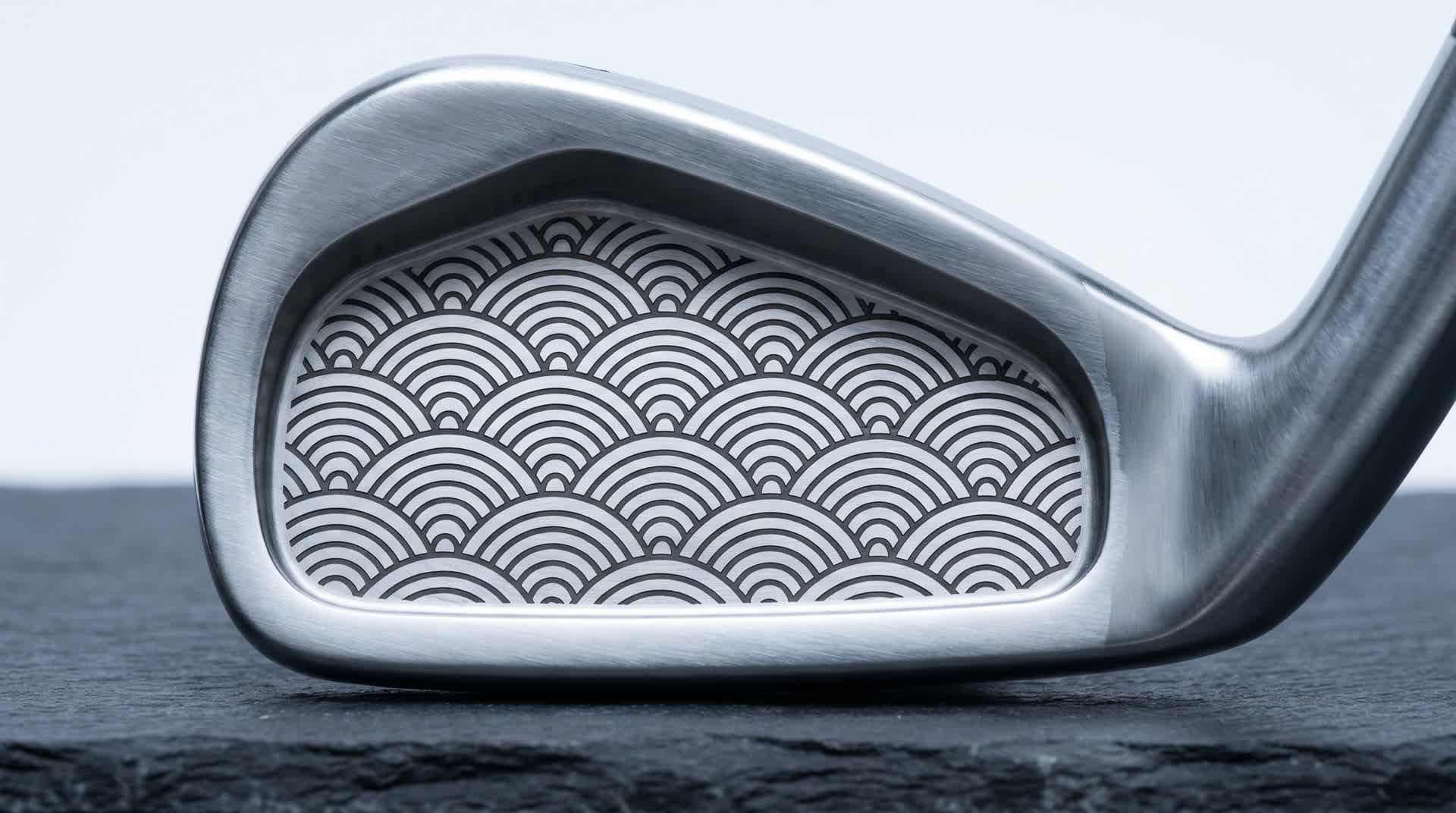 Macro of a premium golf iron cavity laser black-annealed with a repeating Japanese seigaiha wave pattern in fine line-art, flat dark marks on brushed satin steel, shot on dark slate in cool studio light